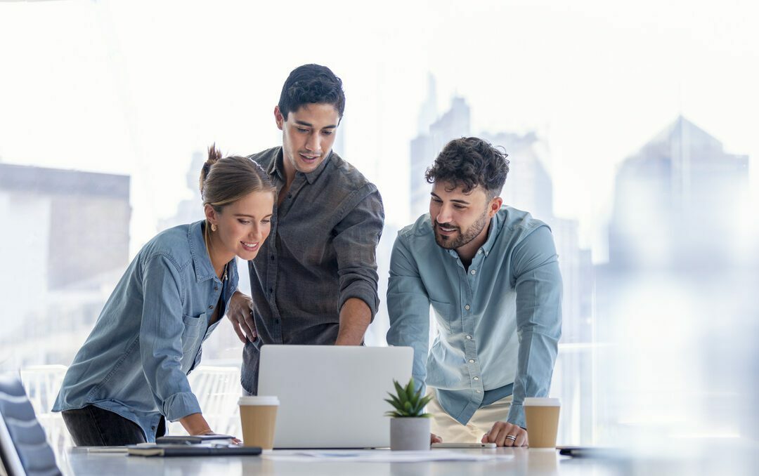 Business team working on a laptop computer. Three people are wearing casual clothing. They are standing in a board room. Multi ethnic group with Caucasian and Latino men and women